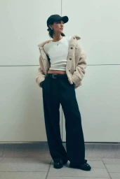 Model wearing a black cap, white top, sherpa jacket, and wide trousers against a pale tiled wall.