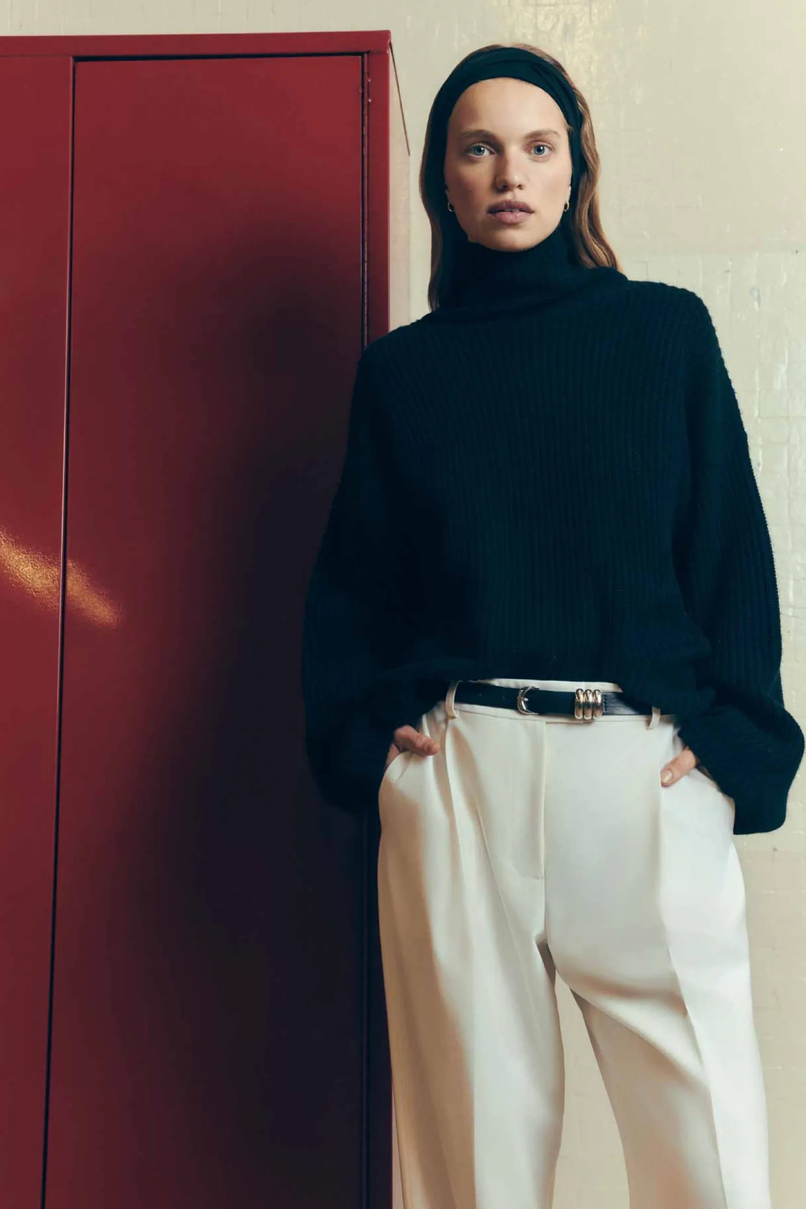 Fashion shot of a woman in a black turtleneck sweater and white pants, posing in front of a red panel. She has a black headband.