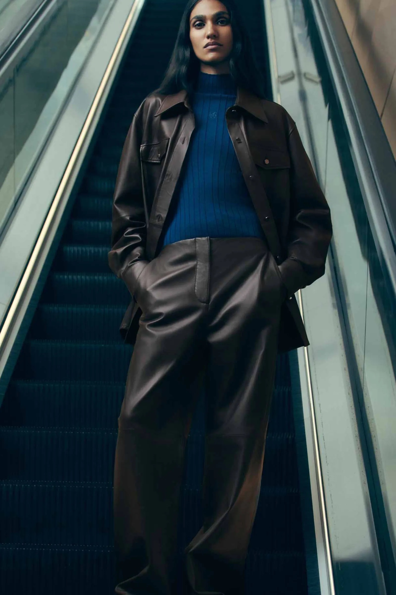 Fashionable woman in leather jacket and pants posing on escalator with hands in pockets, wearing blue ribbed turtleneck top, dark hair.