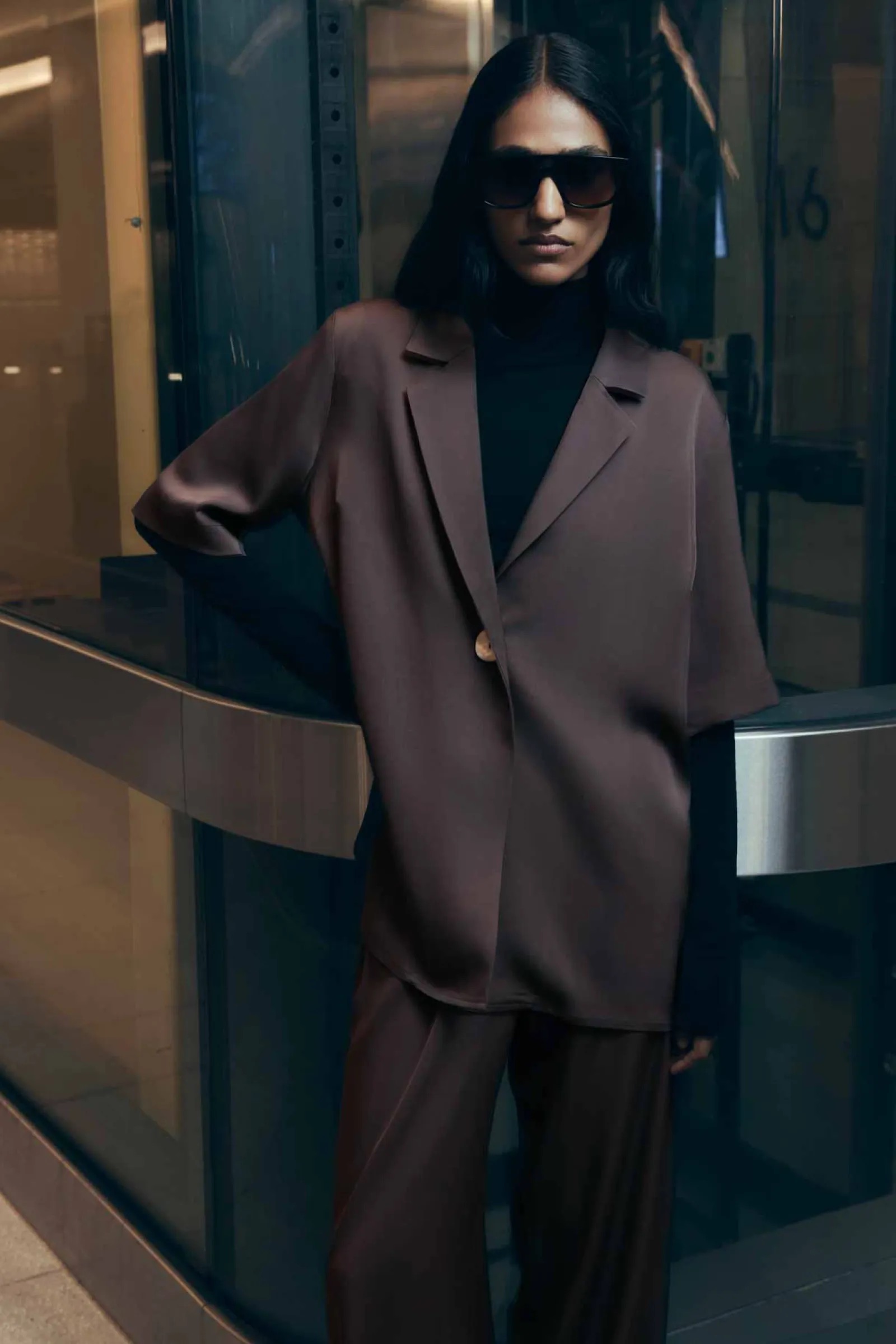 Model in dark sunglasses wearing a brown blazer and trousers standing against a glass building exterior