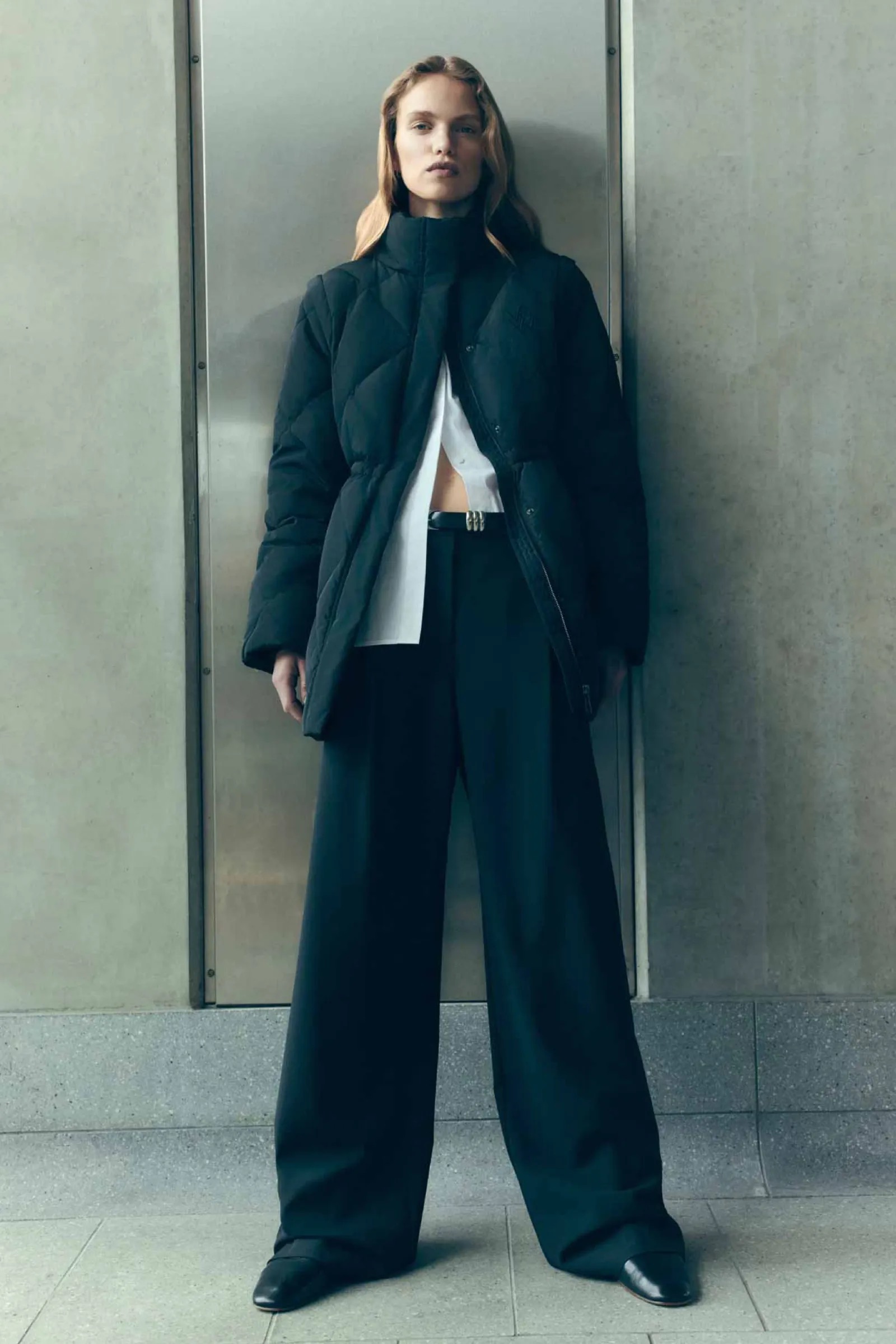A woman in a black puffer coat and wide-leg pants stands against a neutral wall in a full shot, showcasing a modern fashion style.