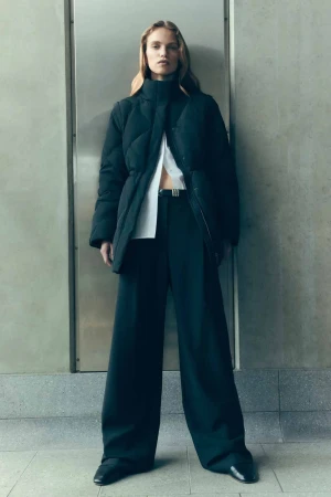 A woman in a black puffer coat and wide-leg pants stands against a neutral wall in a full shot, showcasing a modern fashion style.