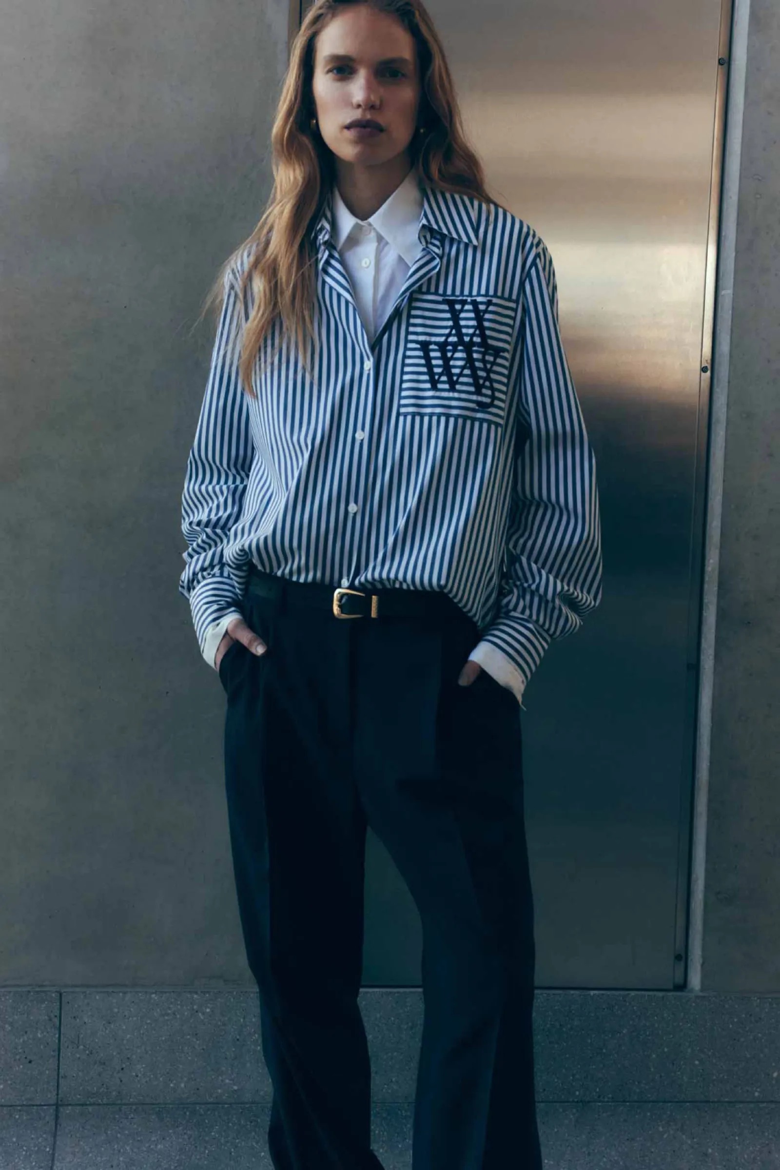 A woman poses indoors wearing a striped button-down shirt and black pants with a gold buckle, creating a fashion-forward editorial shot.