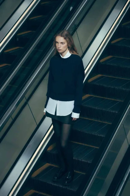 A fashion shot of a young adult woman with long blonde hair on an escalator wearing a black sweater, white shirt, and black tights.
