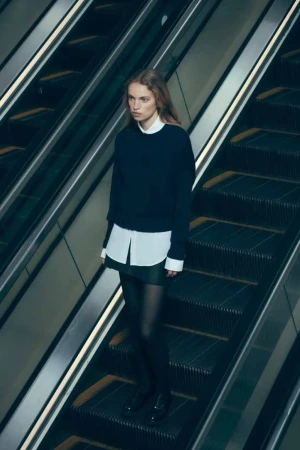 A fashion shot of a young adult woman with long blonde hair on an escalator wearing a black sweater, white shirt, and black tights.