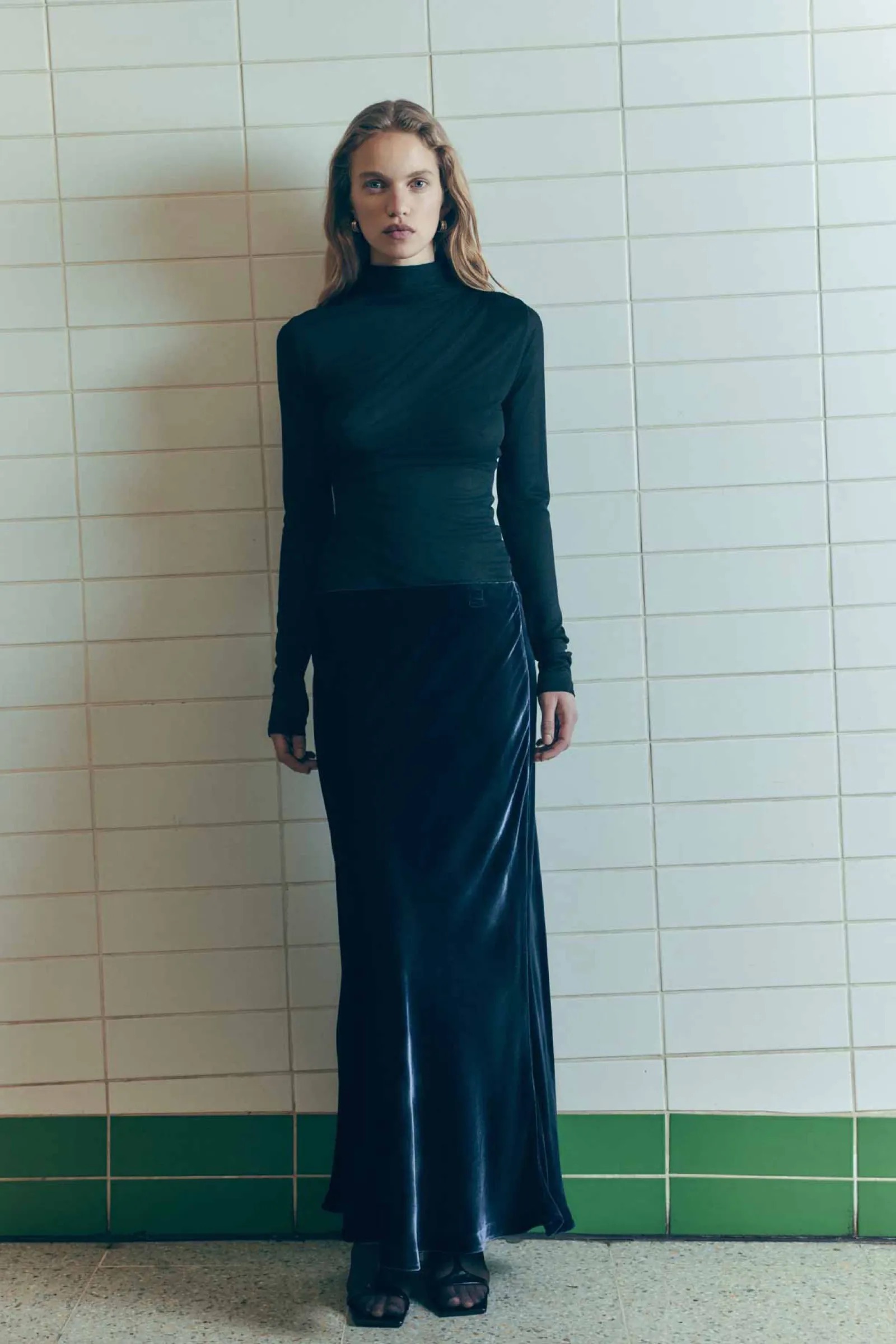Full-length shot of a woman in a dark long-sleeve top and velvet skirt, posing against a neutral-toned tiled wall with green trim.