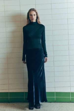 Full-length shot of a woman in a dark long-sleeve top and velvet skirt, posing against a neutral-toned tiled wall with green trim.