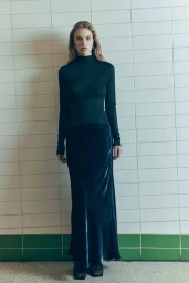 Model standing against white tiled wall wearing dark turtleneck and long velvet skirt for Viktoria & Woods Iridescence project.