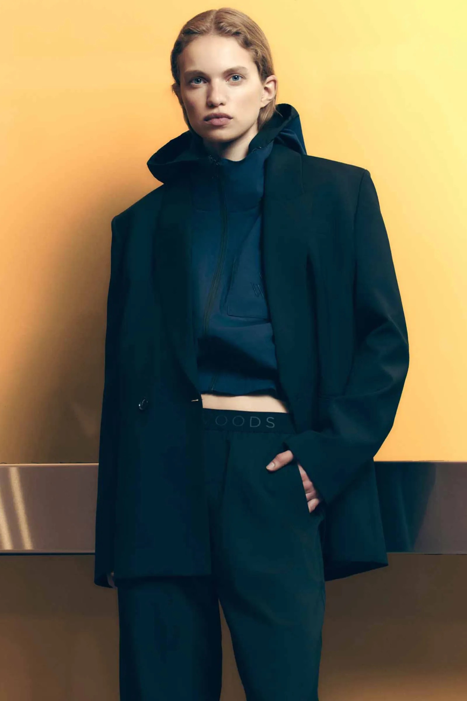 Woman poses indoors against a yellow wall wearing a black blazer, navy hoodie, and trousers in a fashion-style portrait.