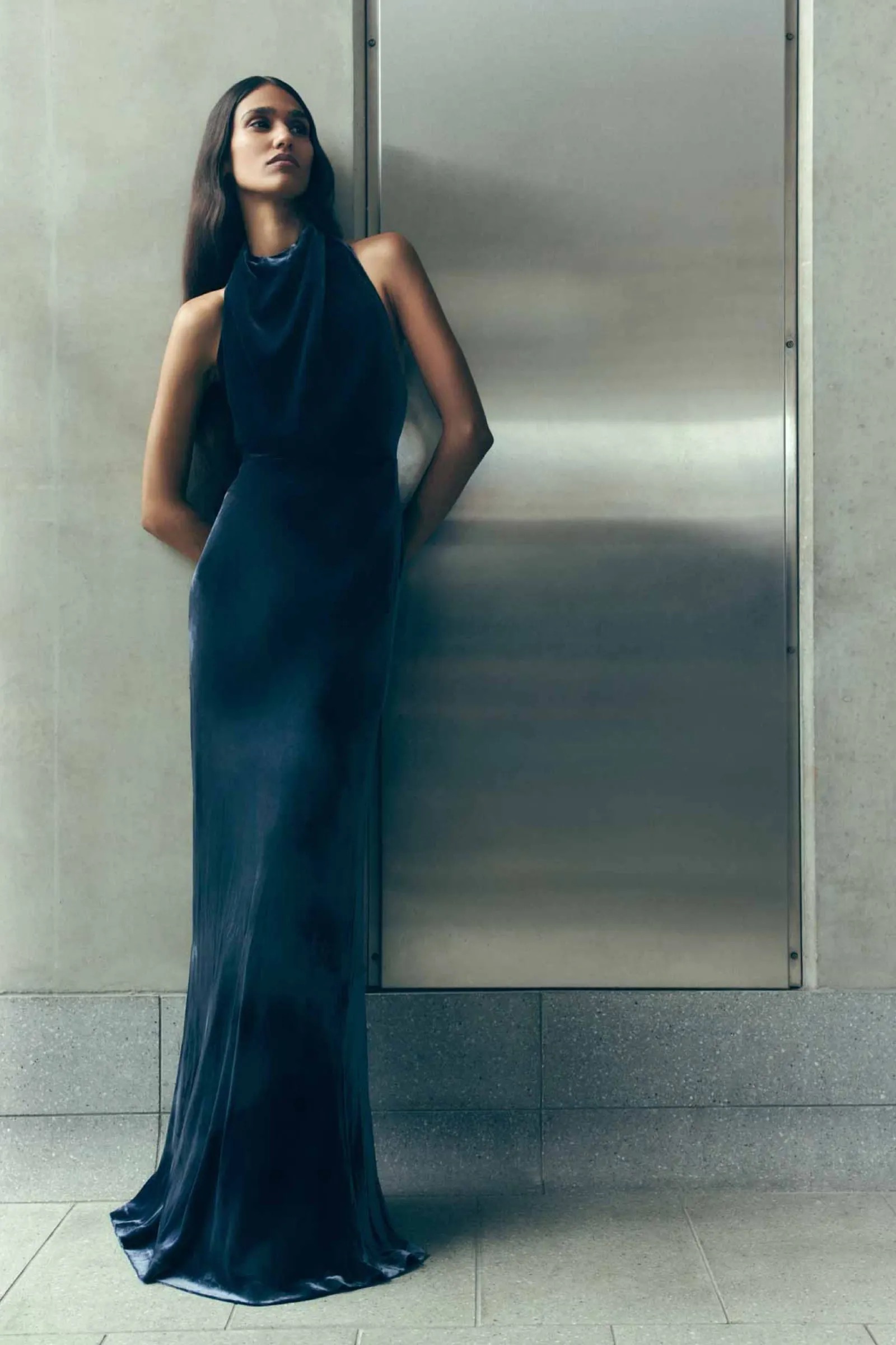 Elegant woman in a long blue velvet dress leaning against a wall. High fashion photography featuring soft, diffused lighting in an indoor setting.