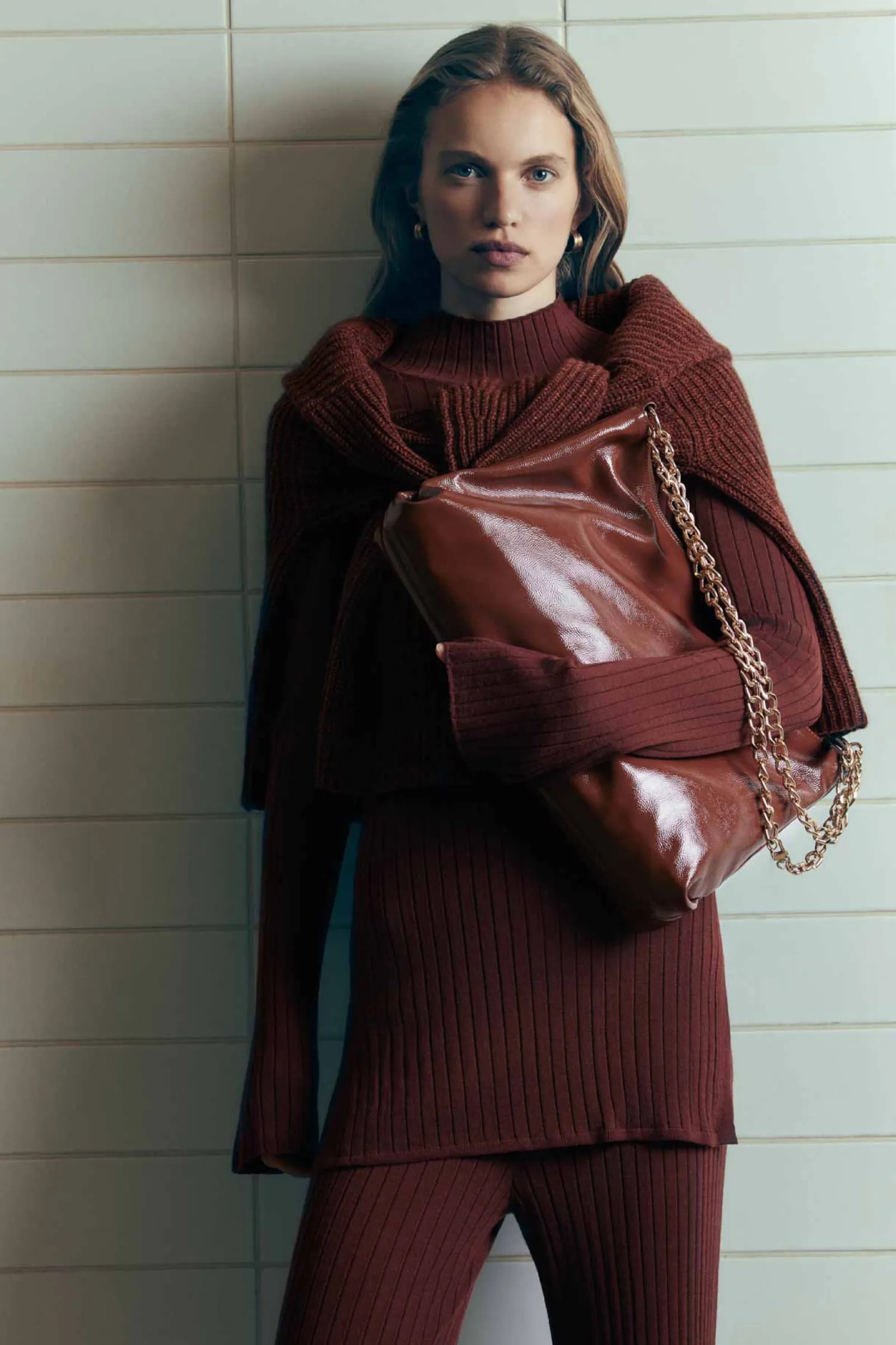 A fair-skinned woman poses indoors, wearing a matching brown sweater and pants, accessorized with a brown purse with a chain strap.