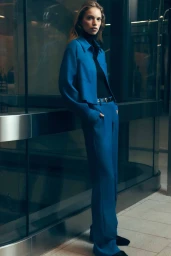 Model in a bright blue suit jacket and matching trousers leaning against a glass wall with dark metal framing.