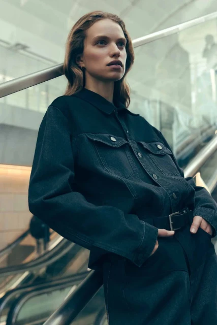 A long-haired woman models black denim shirt and pants on grey escalator, with shallow depth of field and high angle of view.