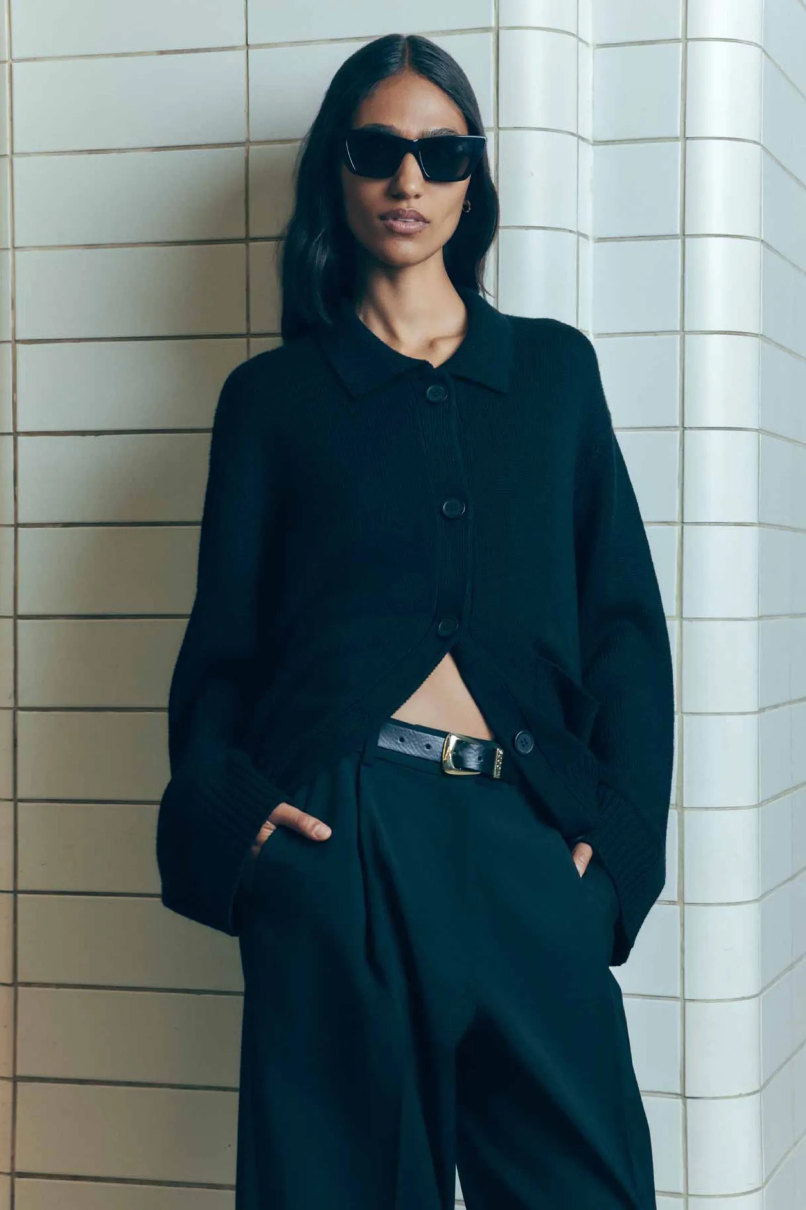 Woman in black sunglasses, cardigan, and trousers poses against a tiled wall, showcasing a blend of fashion and minimalist style.