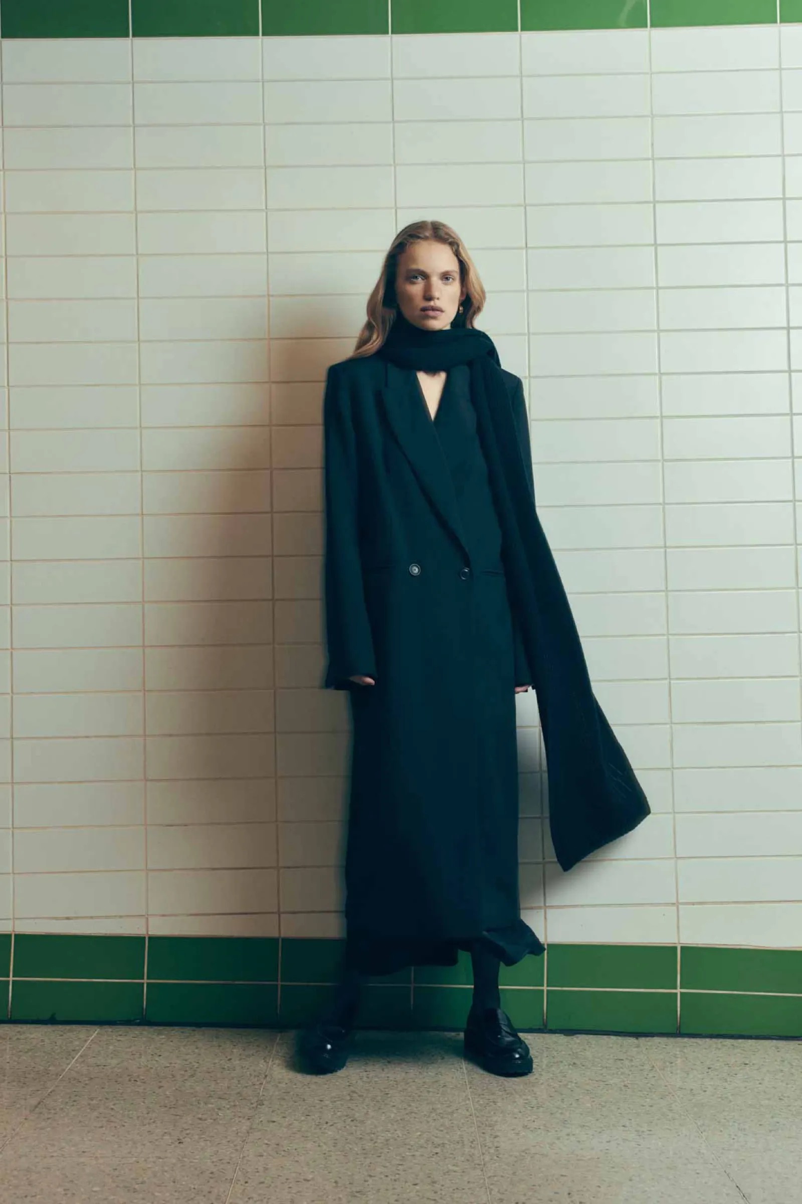 A slender woman posing against a tiled wall, wearing a long dark coat, scarf, and shiny shoes, captured in a full shot.