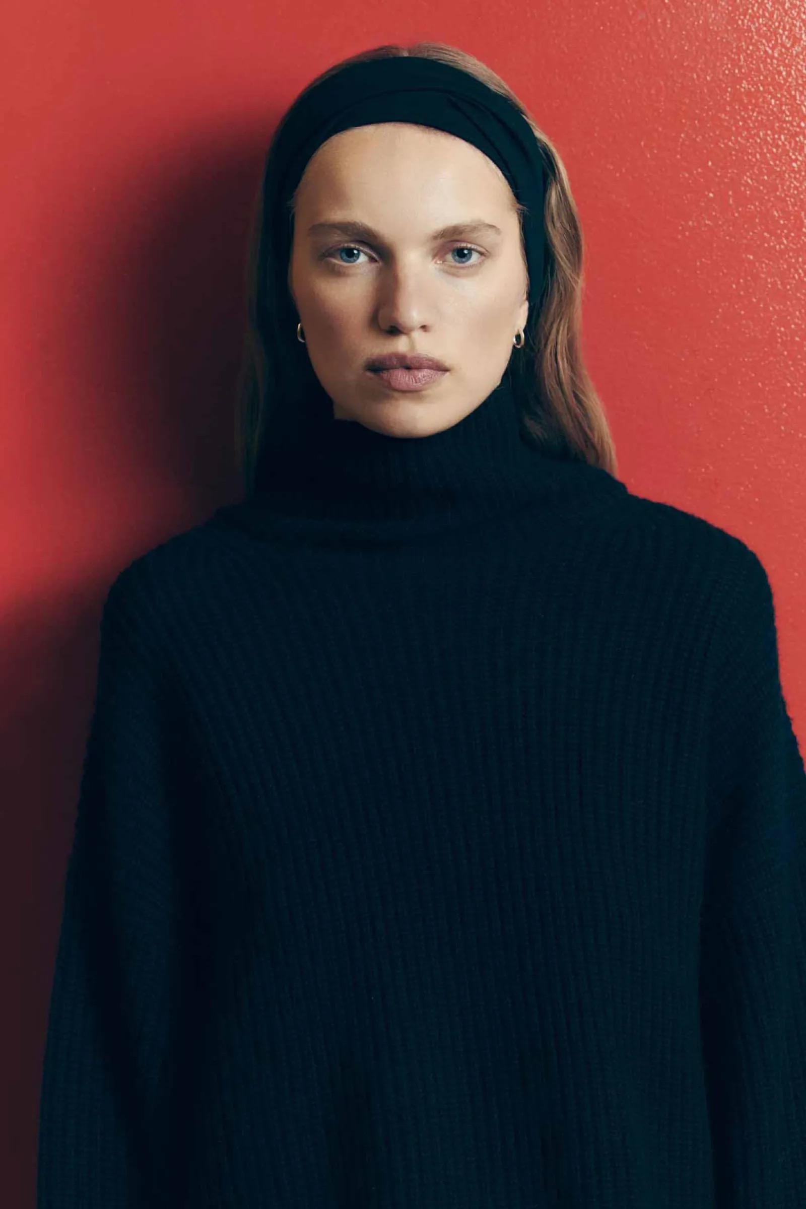 Portrait of a fair-skinned woman with long light brown hair wearing a black turtleneck sweater and headband against a red background.