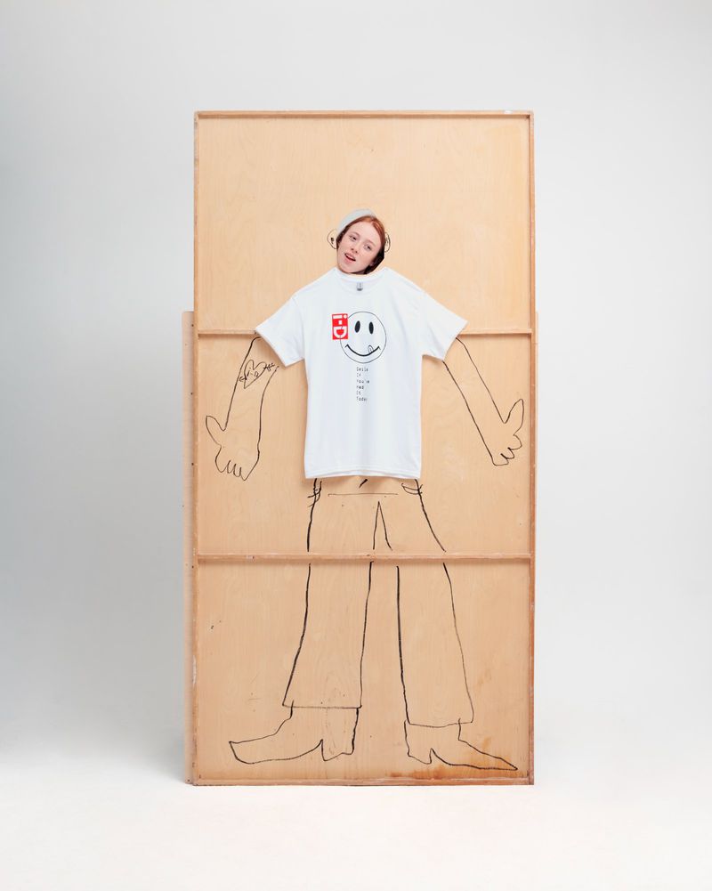 An adult woman peeks through a hole in a wooden frame decorated with a drawing of a human body. She wears a white t-shirt.