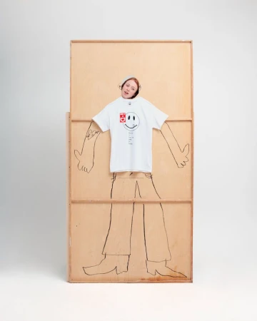 An adult woman peeks through a hole in a wooden frame decorated with a drawing of a human body. She wears a white t-shirt.