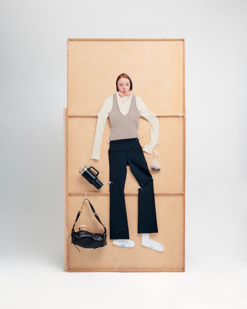 Fashion shoot featuring a model with red hair wearing a beige top and black pants, styled against a tan wooden backdrop.