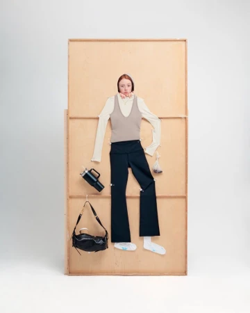 Fashion shoot featuring a model with red hair wearing a beige top and black pants, styled against a tan wooden backdrop.
