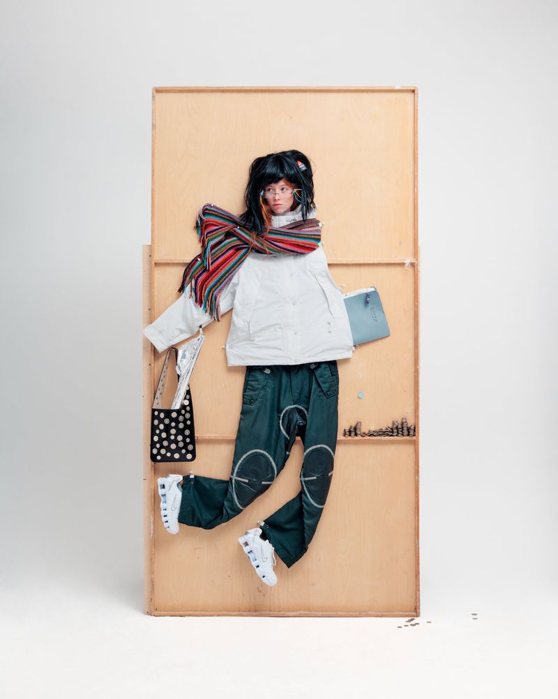 A person in an oversized white jacket and dark pants stands against a beige panel holding a small blue purse and a polka-dot bag.