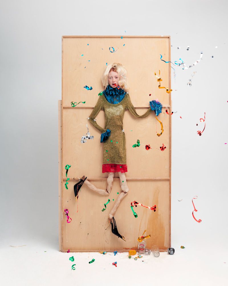 Blonde adult female fashion model stands on painted backdrop amid floating confetti in a studio, wearing an olive-green dress and blue accessories.
