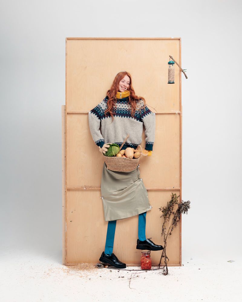 A red-haired woman in a sweater holding a basket of vegetables poses in front of a plywood structure in a studio with a jar of preserves.