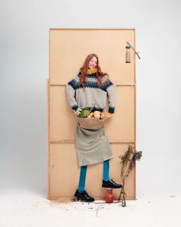 A red-haired woman in a sweater holding a basket of vegetables poses in front of a plywood structure in a studio with a jar of preserves.
