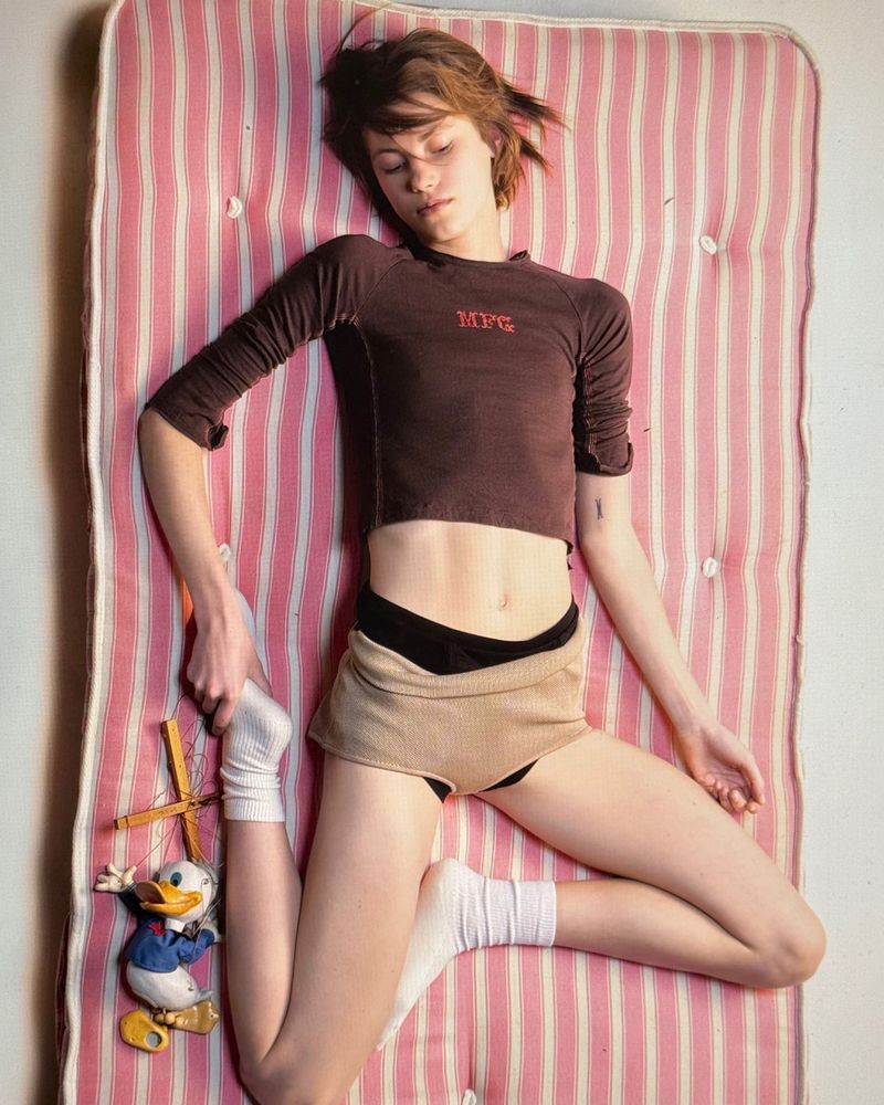 Young adult lying on a pink striped mattress wearing a brown top and shorts with a Donald Duck puppet beside the figure.