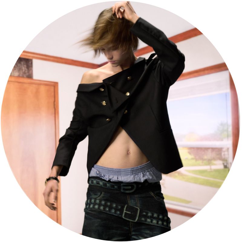 Male fashion model poses in black jacket and jeans in an indoor setting with light hair, accessorized with bracelets and belts.