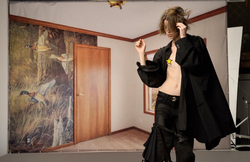 Woman in black shirt and jeans poses in front of a backdrop depicting an interior scene with a door and wildlife painting.