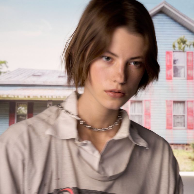 Portrait of a person with short brown hair and a silver chain necklace, posing outdoors in front of a house.