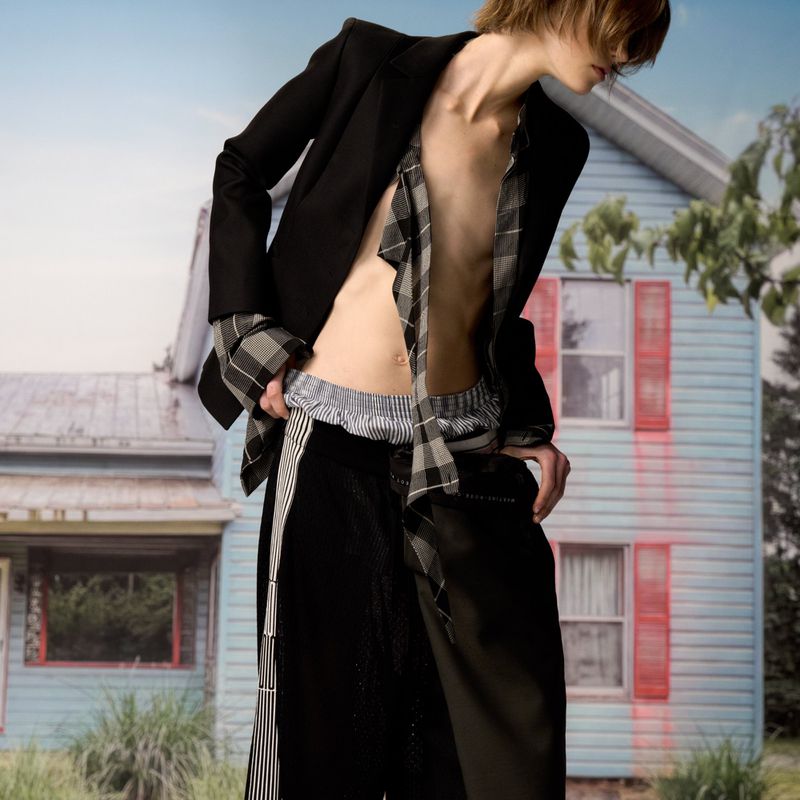 Editorial fashion shot of a slim adult in a deconstructed black blazer and trousers, posing outdoors in front of a light blue house.