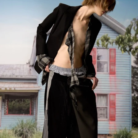 Editorial fashion shot of a slim adult in a deconstructed black blazer and trousers, posing outdoors in front of a light blue house.