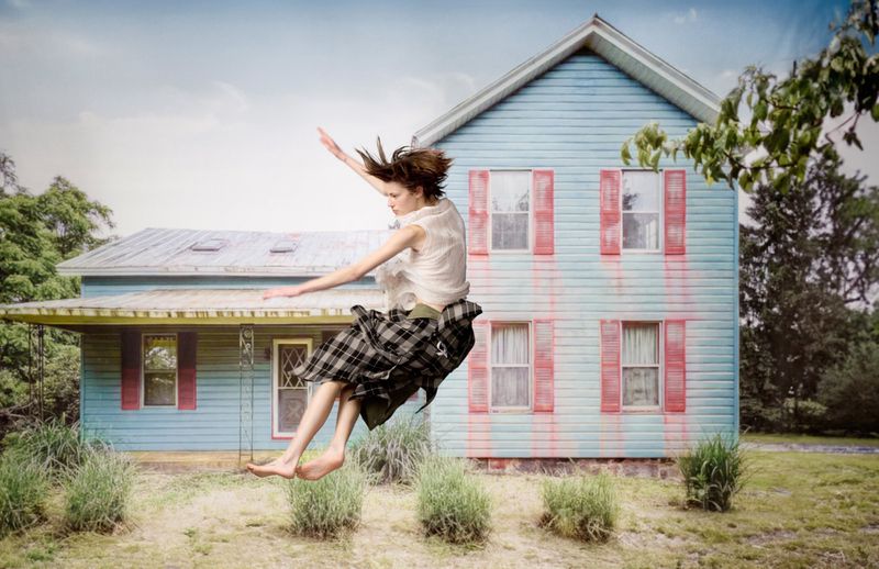 Adult female barefoot and airborne in a plaid skirt and white blouse, captured outdoors with a blue house and grassy field backdrop.
