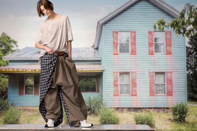 A slender adult poses outdoors wearing a tattered beige t-shirt and deconstructed trousers against a painted house backdrop.