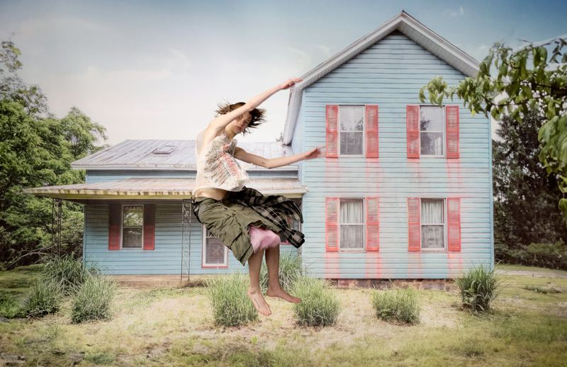 Adult woman in mid-air pose with outstretched arms and legs, stands out against a blue house and partly cloudy sky in a rural setting.