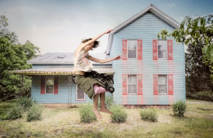 Adult woman in mid-air pose with outstretched arms and legs, stands out against a blue house and partly cloudy sky in a rural setting.