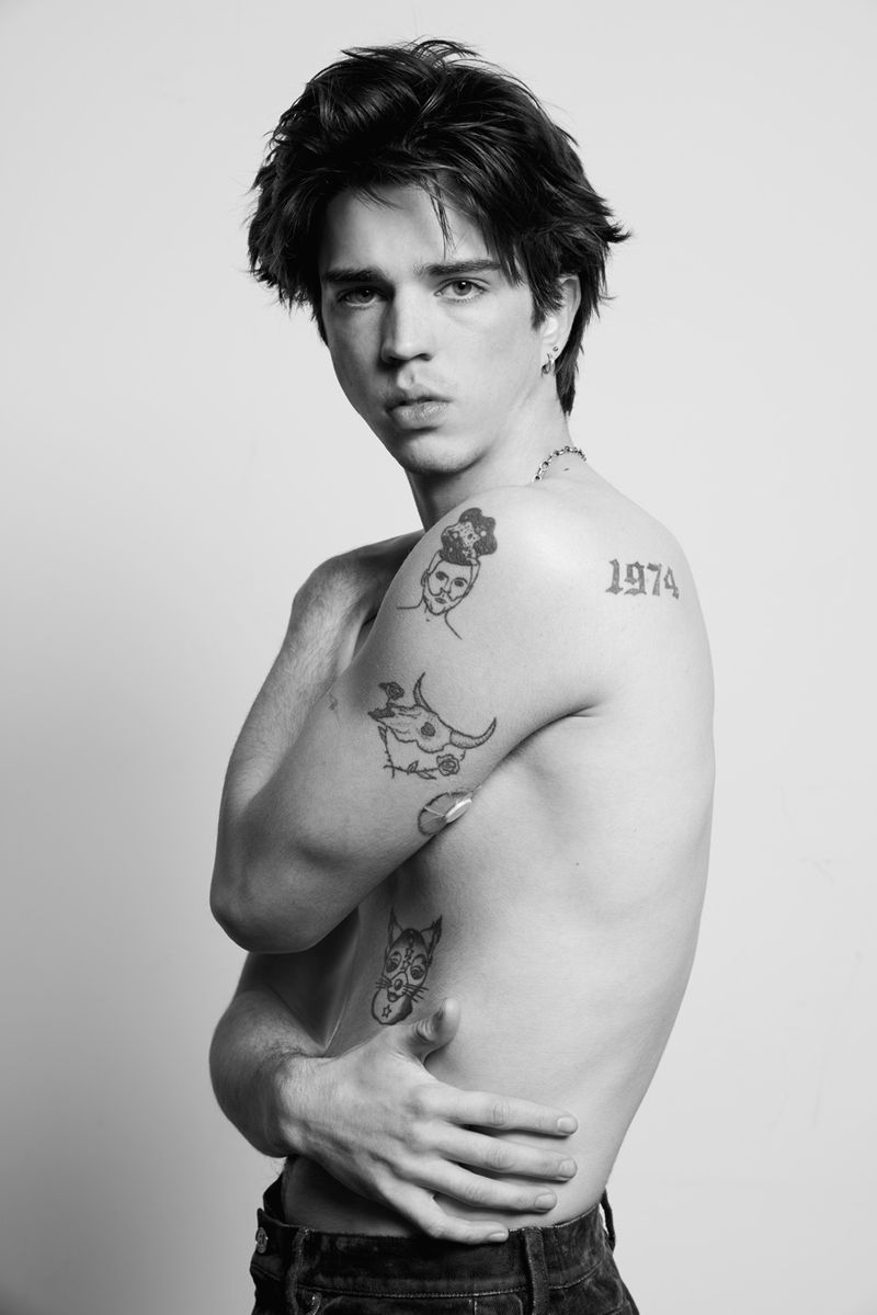 Black and white portrait of a shirtless male model with dark hair and tattoos on his arms and torso, standing in a photo studio.