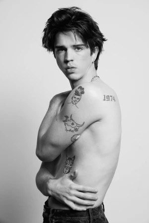 Black and white portrait of a shirtless male model with dark hair and tattoos on his arms and torso, standing in a photo studio.