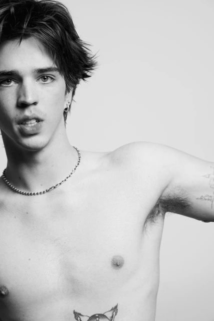 Black and white studio headshot of a bare-chested young adult male with dark hair, silver necklace, and tattoos against a plain background.