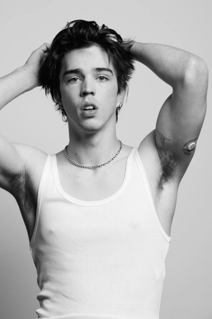 Monochrome portrait of a young adult male with dark hair, wearing a white tank top, posing with his hands behind his head.