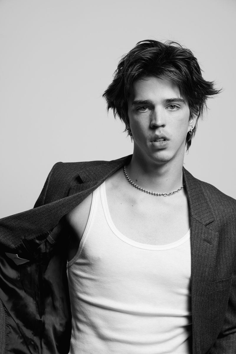 Monochrome portrait of a young adult male in an open dark grey jacket and white tank top, posing in a photo studio setting.