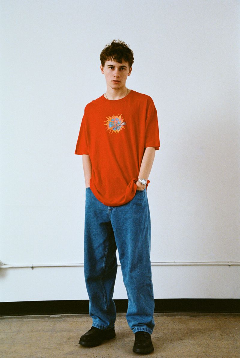 A young adult male stands in a photo studio wearing a red t-shirt with a graphic, blue jeans, and black shoes against a white backdrop.