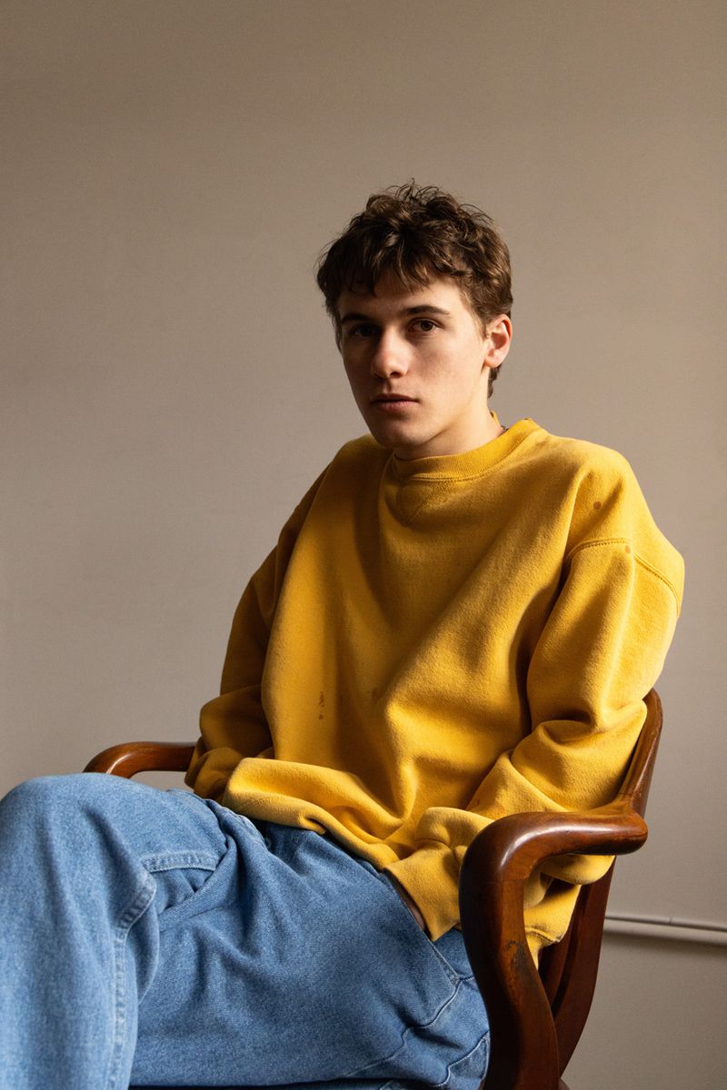 Sam Nivola seated in a wooden armchair wearing a mustard yellow sweatshirt and blue jeans for HERO Magazine.