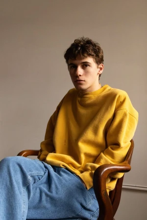 Sam Nivola seated in a wooden armchair wearing a mustard yellow sweatshirt and blue jeans for HERO Magazine.