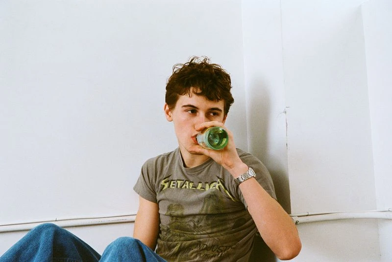 Young adult male in gray t-shirt drinking from green glass bottle indoors against white wall, wearing wristwatch, blue jeans.