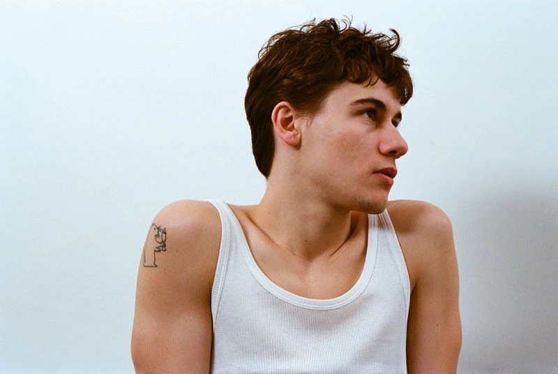 Portrait of a young man with curly dark hair, wearing a white tank top against a white backdrop, gazing off to the side.