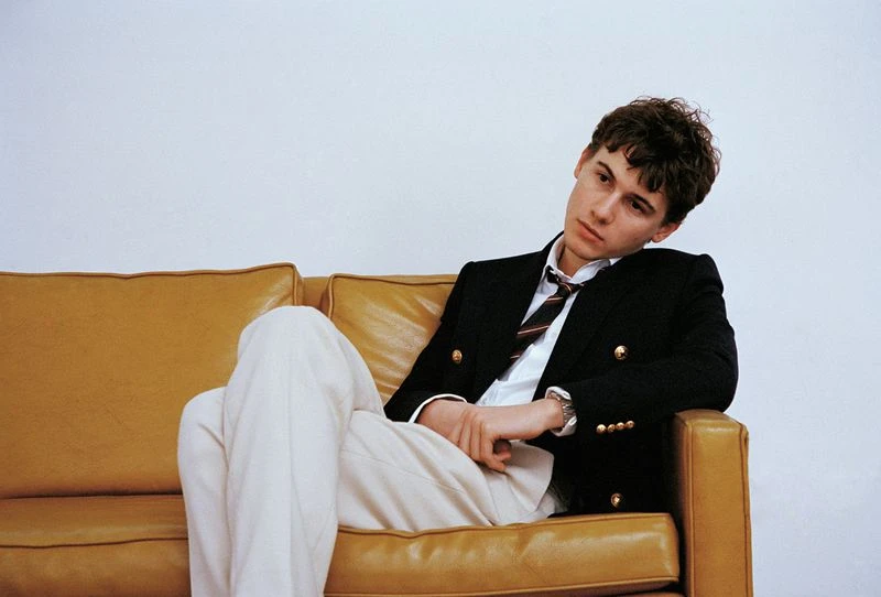 Young adult male in a blazer and slacks poses while reclining on a mustard-colored couch in front of a neutral wall in a studio shot.