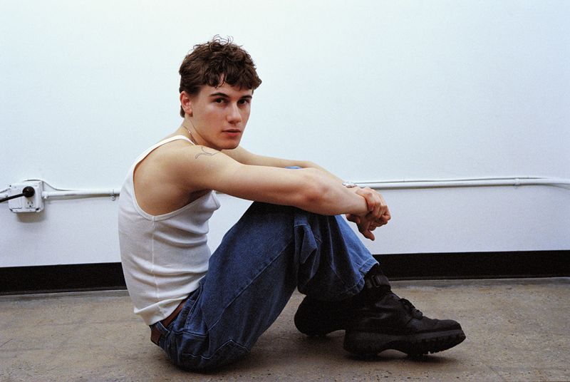 Man in a white tank top and blue jeans sits on the floor in an indoor setting, looking directly at the camera in a medium shot.